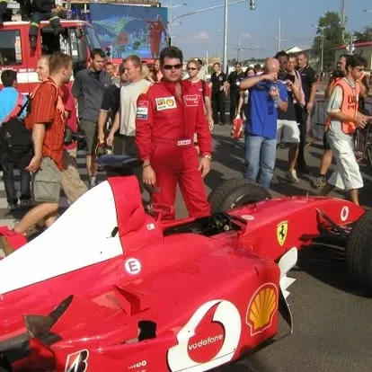 Tom Gryf v červené kombinéze Ferrari vedle vystaveného vozu F1, obklopený davem diváků