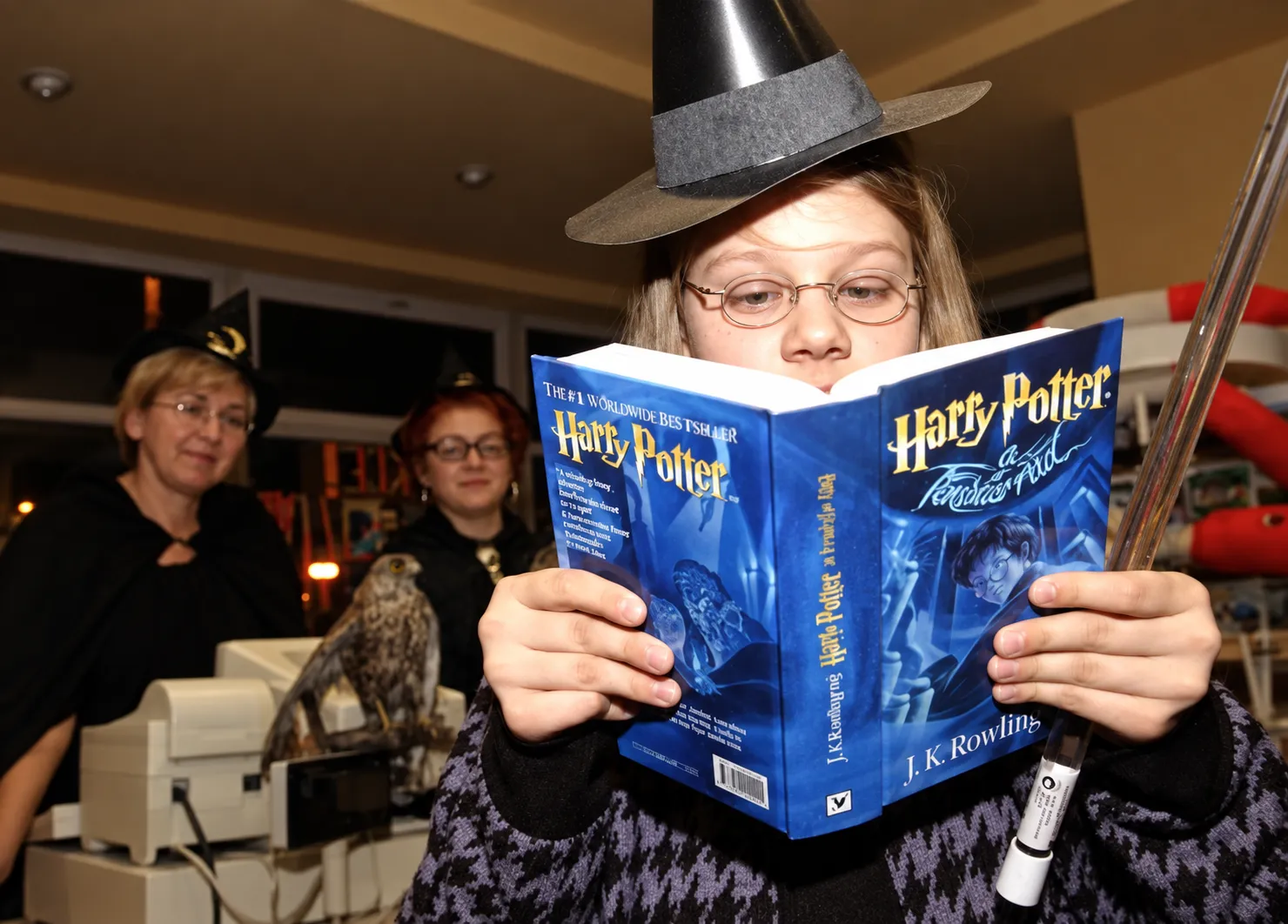 Fanoušek v čarodějnickém klobouku a kulatých brýlích čte české vydání knihy Harry Potter a Fénixův řád, v ruce hůlku; v pozadí kostýmovaný personál a živý sokol na pokladně knihkupectví Kanzelsberger