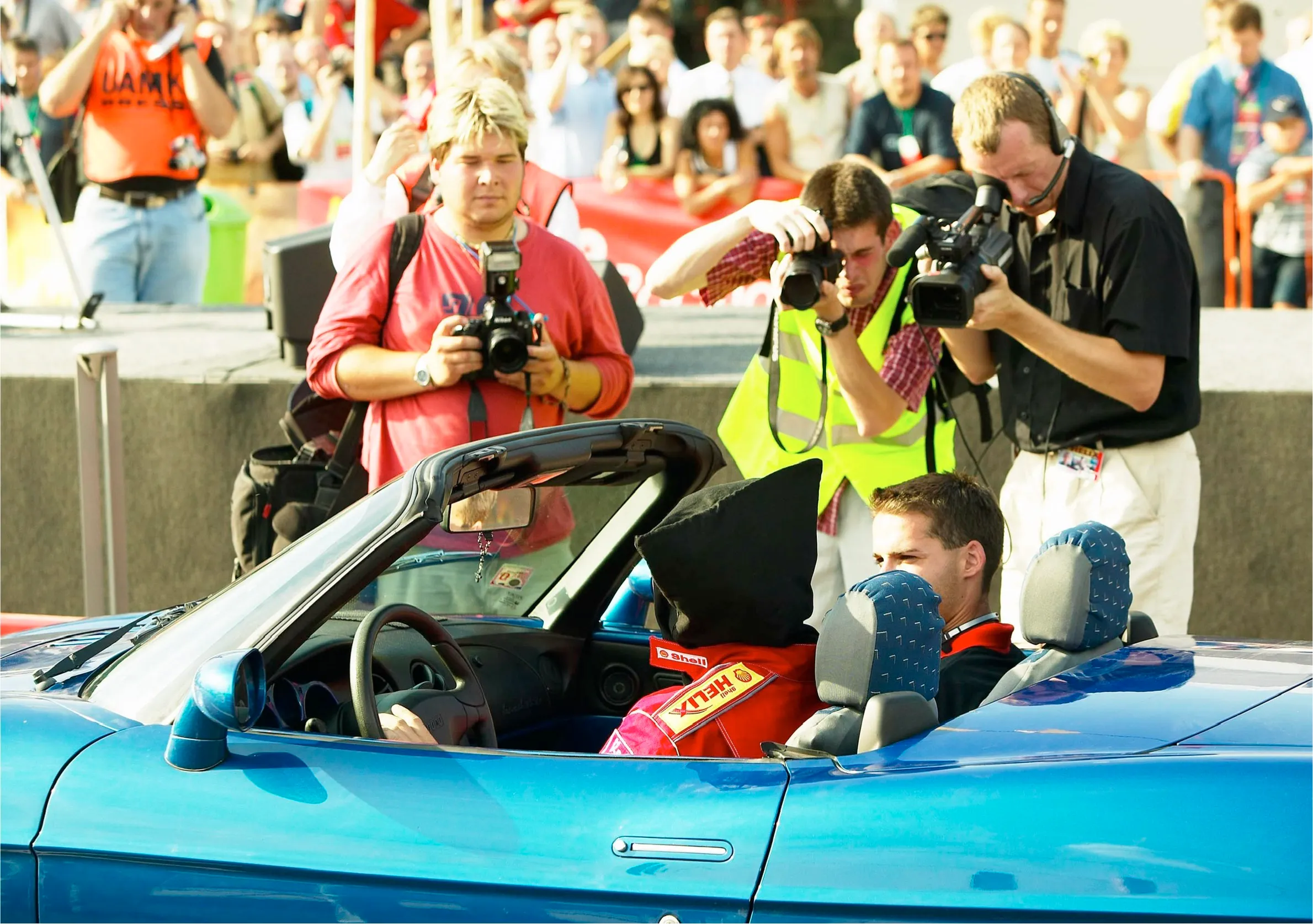 Tom Gryf za volantem modrého kabrioletu Fiat s černou látkovou kuklou přes hlavu, vedle spolujezdec; těsně u vozu skupina fotografů a kameraman, v pozadí plné tribuny diváků