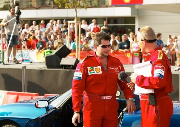 Tom Gryf v červené kombinéze Shell V-Power a slunečních brýlích při rozhovoru s moderátorem, v pozadí diváci a modrý sportovní vůz