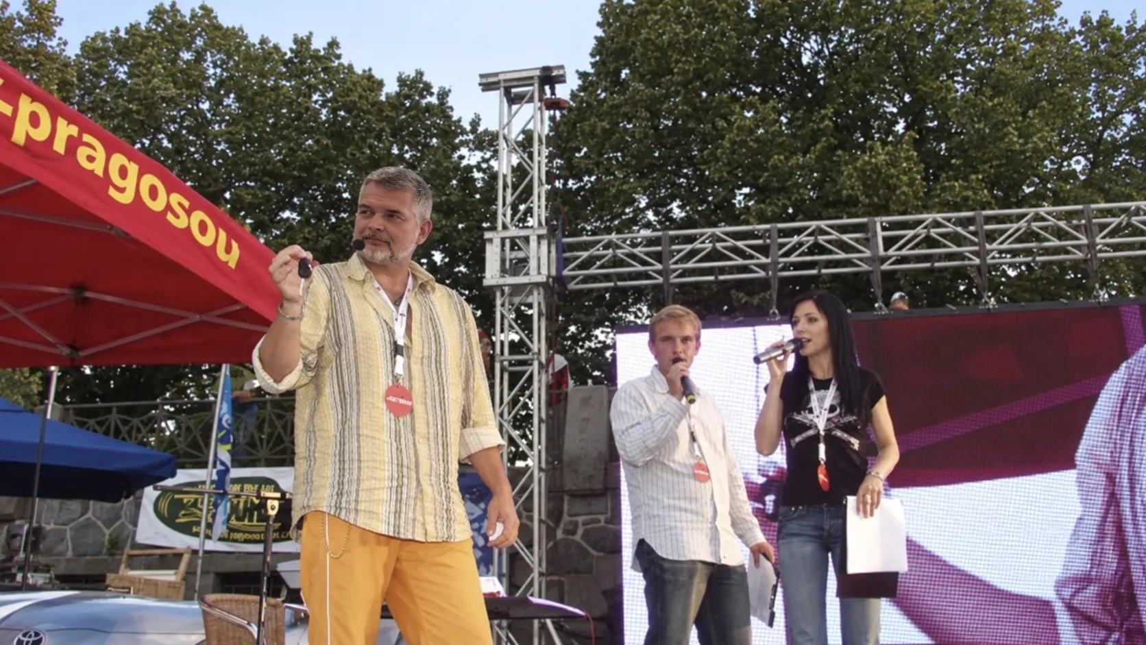 Tom Gryf na venkovním pódiu drží zvednutý klíček od auta, vedle něj dva moderátoři s mikrofony, v popředí automobil