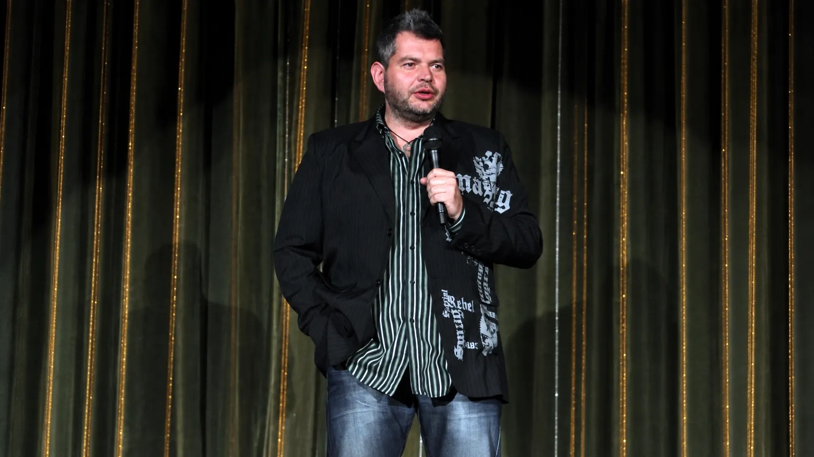 Tom Gryf s mikrofonem na pódiu, v pozadí tmavý závěs se zlatými pruhy